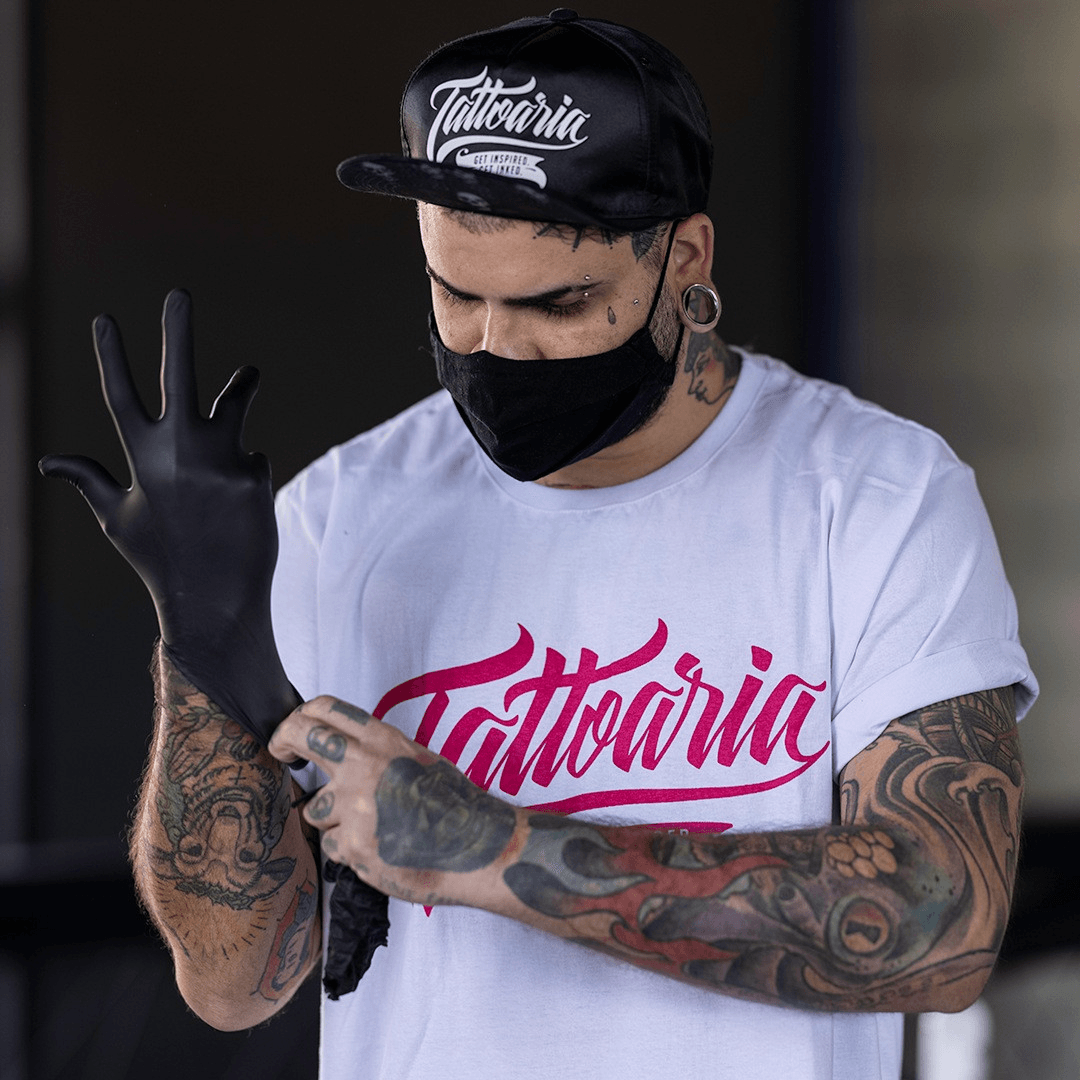 Tatuador convidado de boné e camiseta da Tattoaria calçando a luva preta antes de tatuar.
