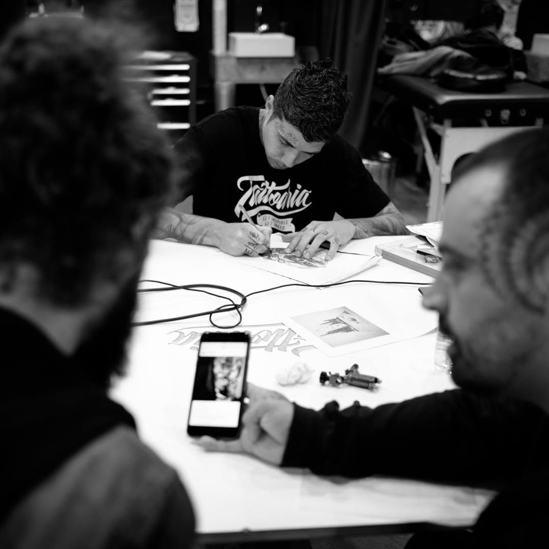 Foto em preto e branco: tatuador de camiseta da Tattoaria desenhando numa mesa, com outros dois em primeiro plano.