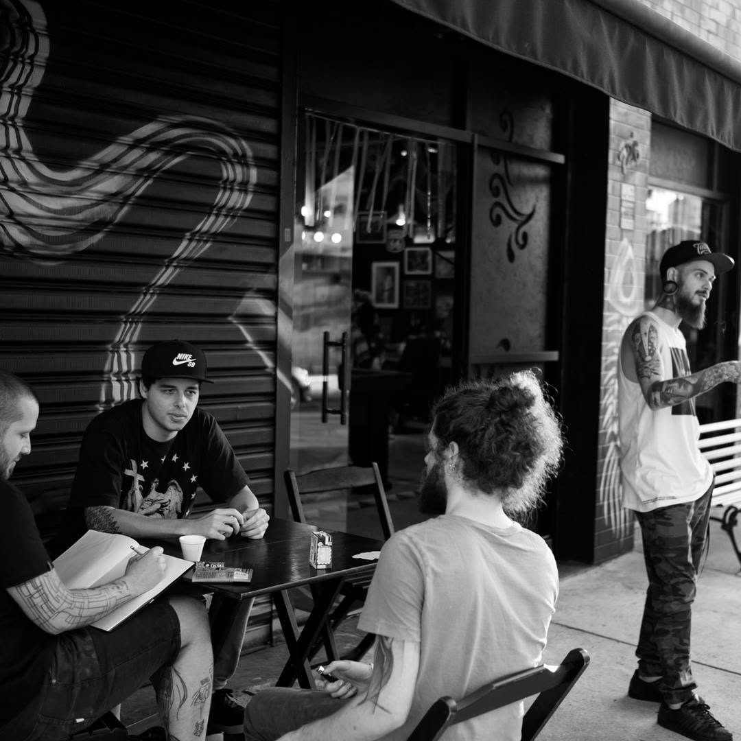 Foto em preto e branco: tatuadores convidados conversando numa mesa na calçada, na porta da Tattoaria.