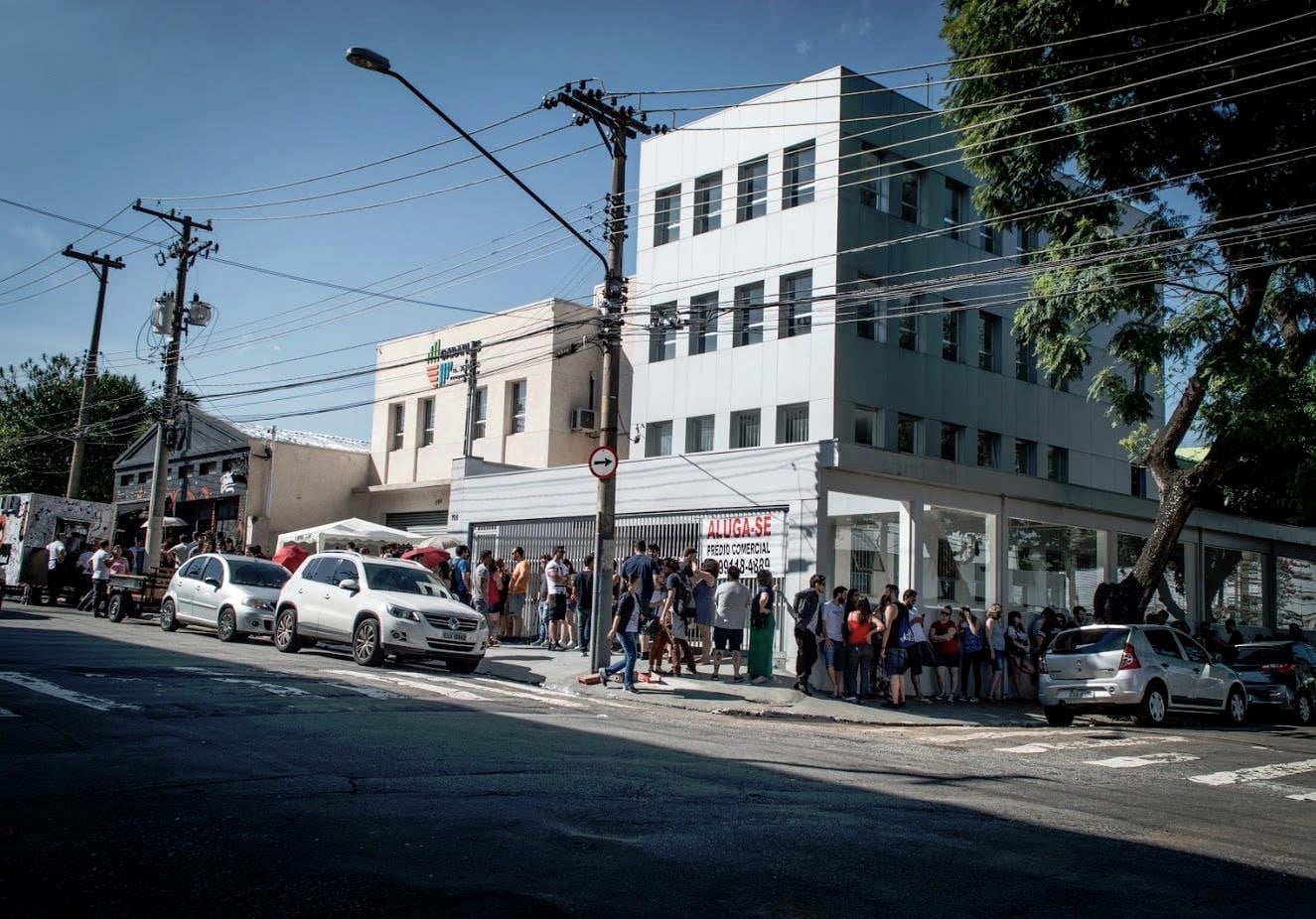 Fila de gente dobrando o quarteirão na porta da Tattoaria num dia de sol, ao longo da calçada.
