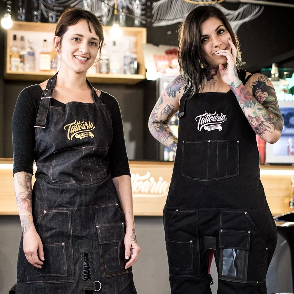 Duas tatuadoras no bar da Tattoaria vestindo os aventais jeans e preto com o logo da marca.