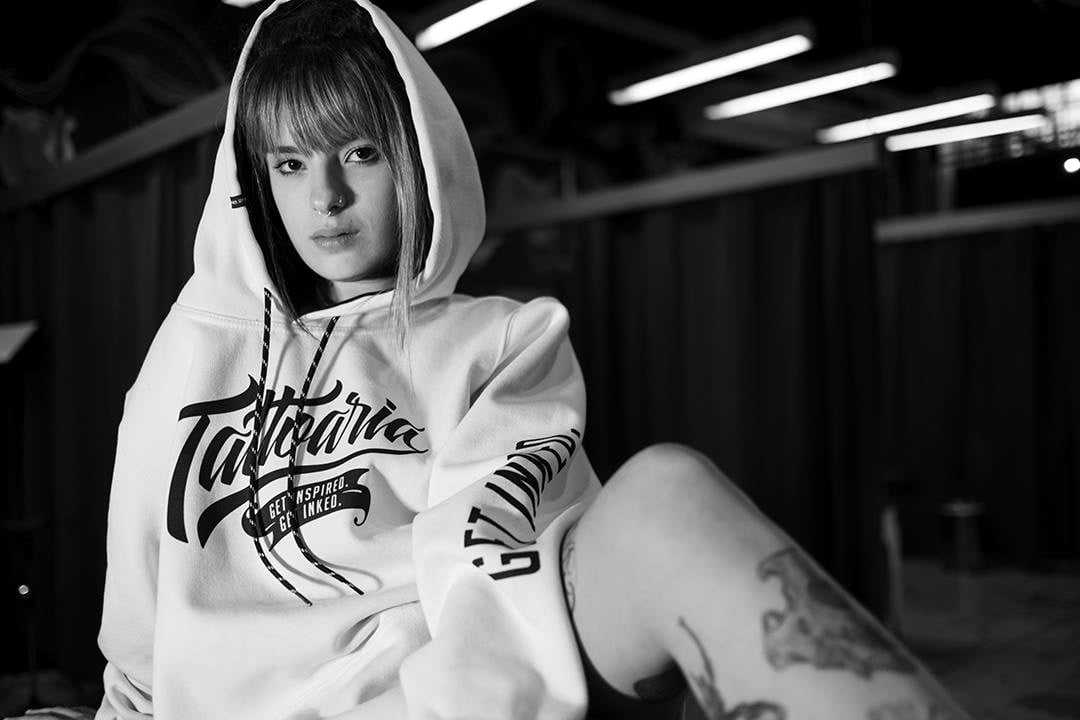 Foto em preto e branco: uma mulher de moletom branco da Tattoaria com capuz, sentada no galpão.
