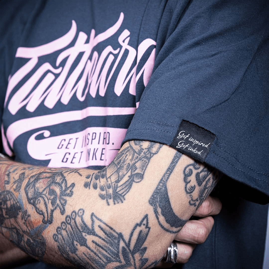 Detalhe da camiseta azul-marinho da Tattoaria com estampa rosa e a etiqueta Get inspired. Get inked. na manga, num braço tatuado.