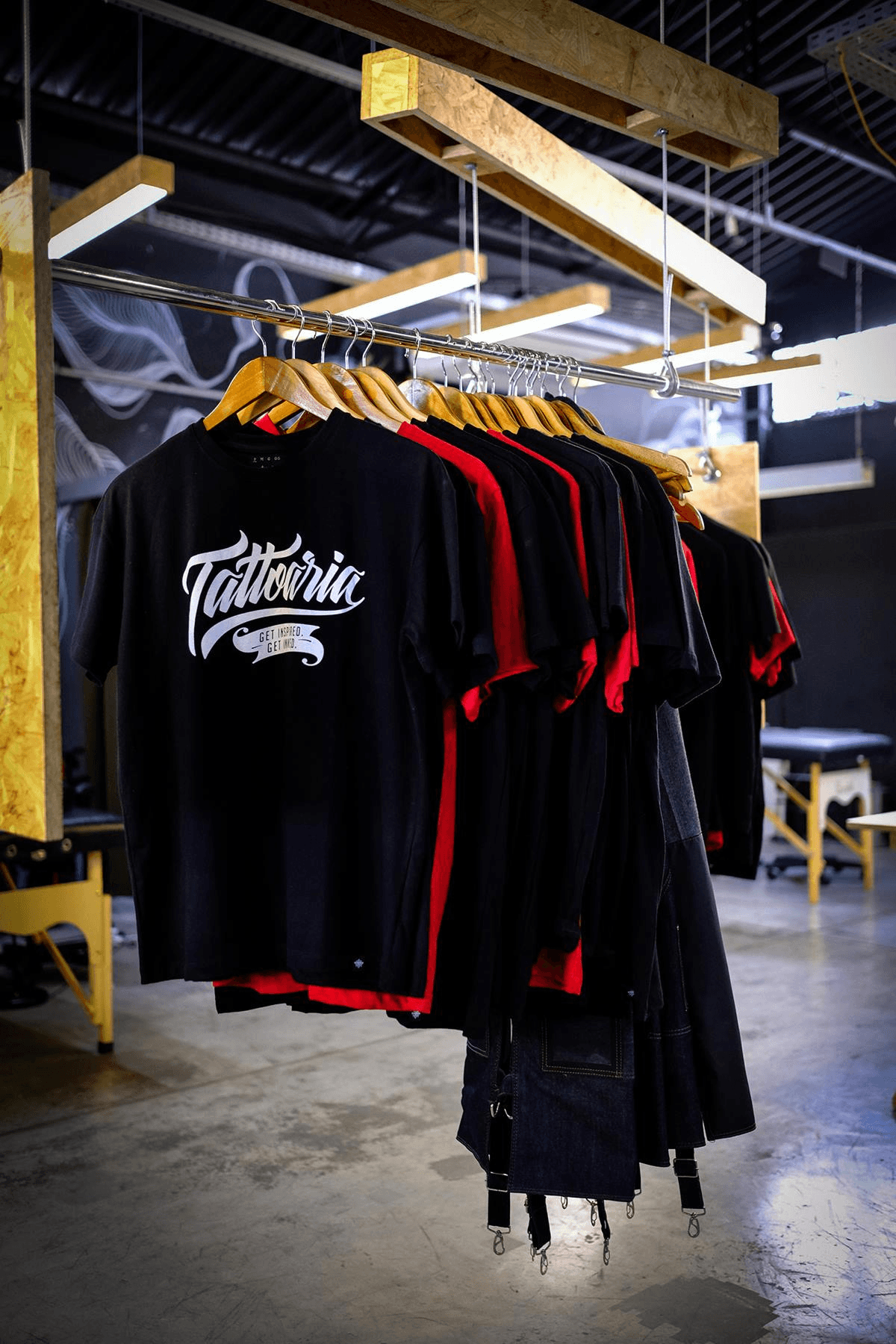 Camisetas pretas e vermelhas da Tattoaria penduradas numa arara de madeira, dentro do galpão.