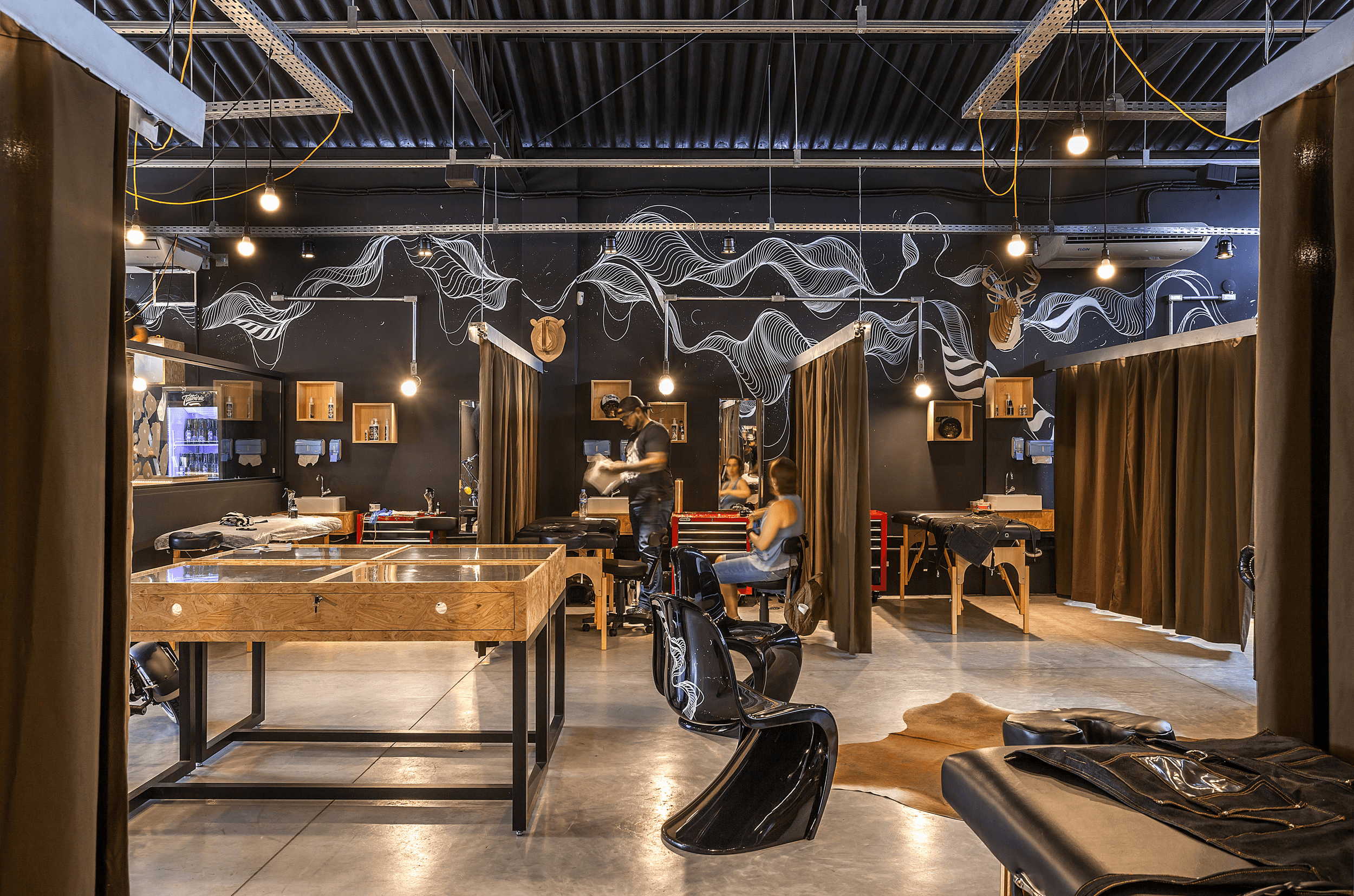 Galpão da Tattoaria: paredes pretas com mural de linhas brancas, baias de tatuagem abertas entre cortinas, vitrine de madeira e um tatuador atendendo uma cliente.